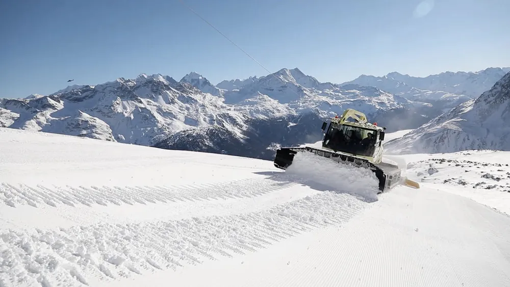 A snow groomer cutting across a slope below snow-covered peaks