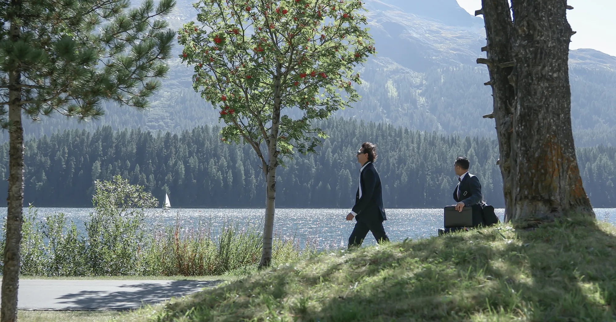 Two men in dark suits walking along a lake shore below a forested mountain