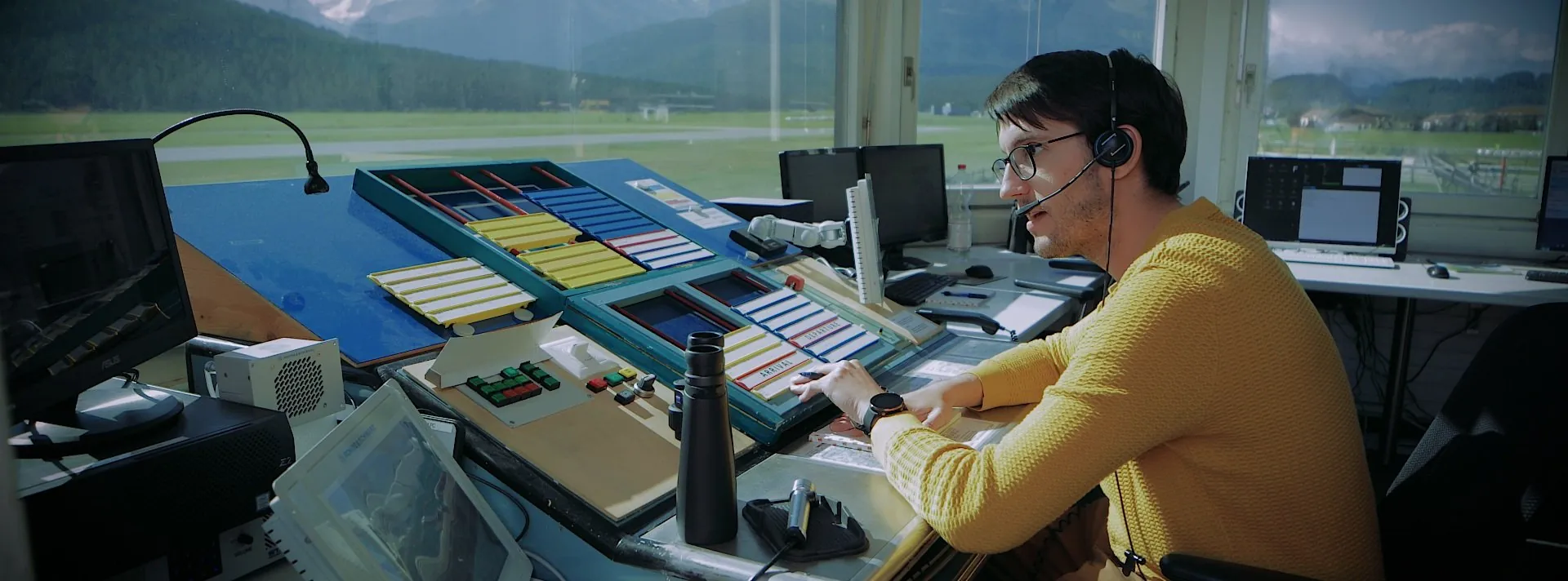 A commentator wearing a headset at a live production desk, overlooking a mountain valley